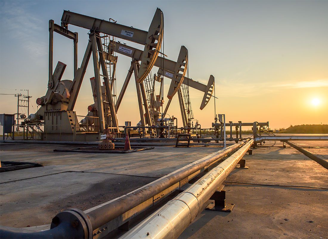 About Our Agency - View of a Row of Oil Drilling Rigs with Surrounding Pipes Along the Ground at Sunset at an Oil Production Facility