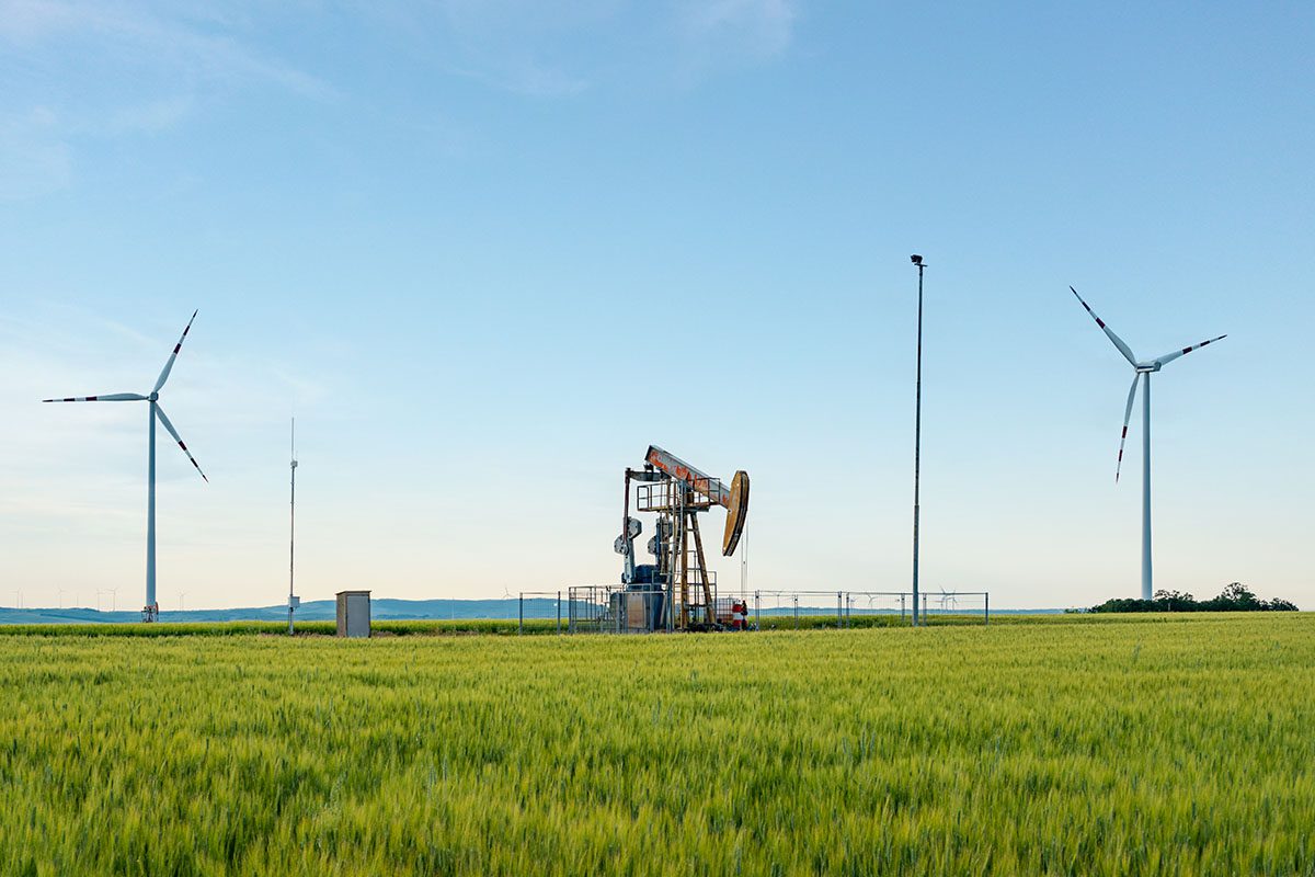 Contact - View of an Oil Drilling Rig on an Open Field of Green Grass with a Wind Turbine on Both Sides