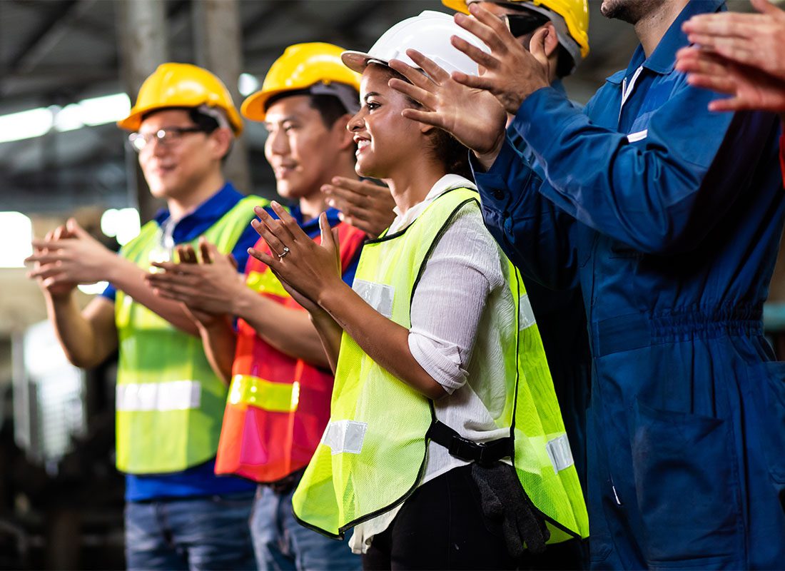 Employee Benefits - Group of Diverse Employees Wearing Hard Hats Clapping Their Hands to Celebrate Success While Standing in a Factory
